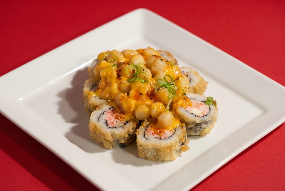 Fried sushi rolls topped with scallops, orange sauce, sesame seeds, and green onions on a white plate.