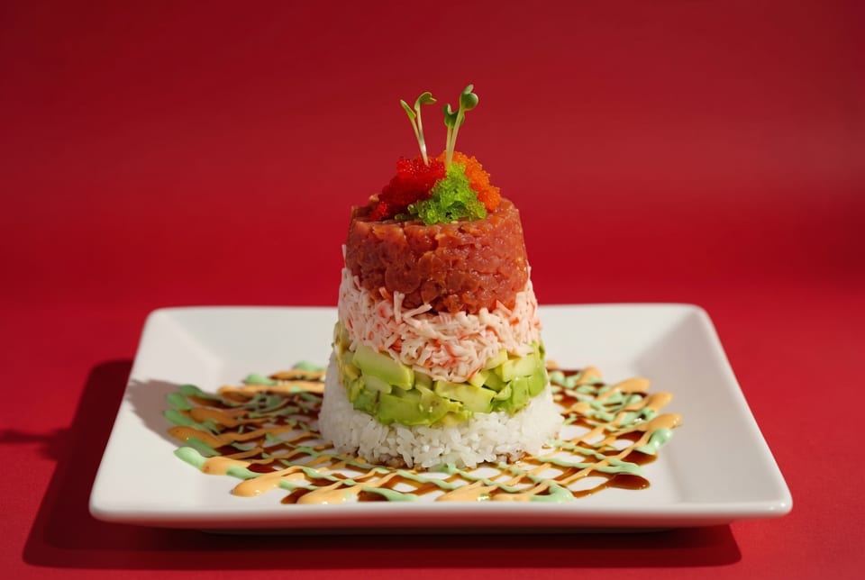 Sushi tower with layers of rice, avocado, crab, tuna, and roe, drizzled with sauces.