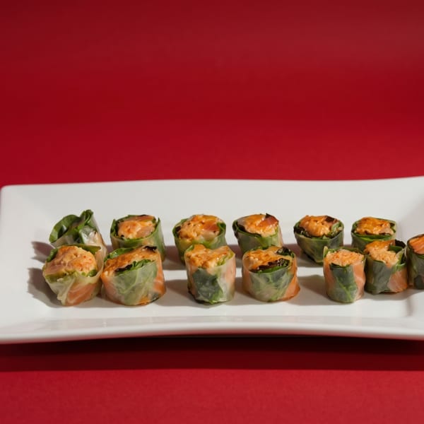 Twelve fresh spring rolls with salmon and greens on a white rectangular plate against a red background.