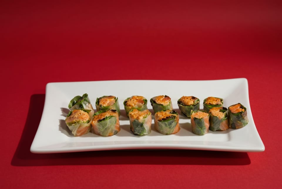 Twelve fresh spring rolls with salmon and greens on a white rectangular plate against a red background.