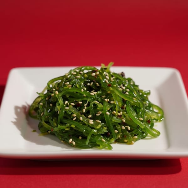 Bright green seaweed salad topped with sesame seeds on a white square plate against a red background.