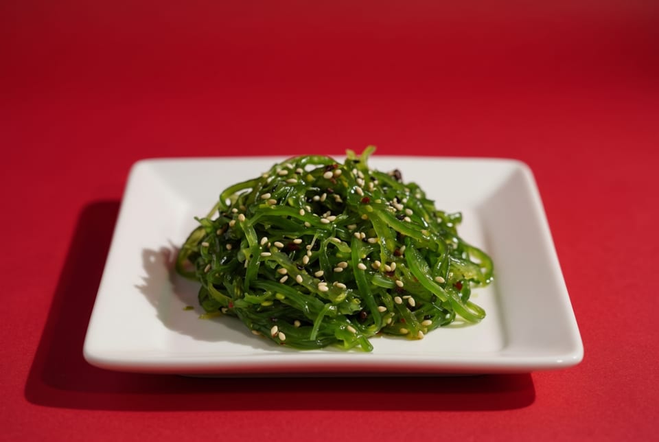 Bright green seaweed salad topped with sesame seeds on a white square plate against a red background.