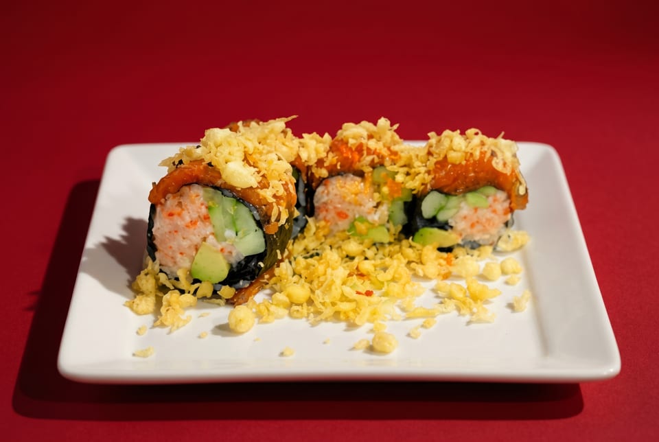 Three sushi rolls with orange sauce and crispy toppings on a white plate against a red background.