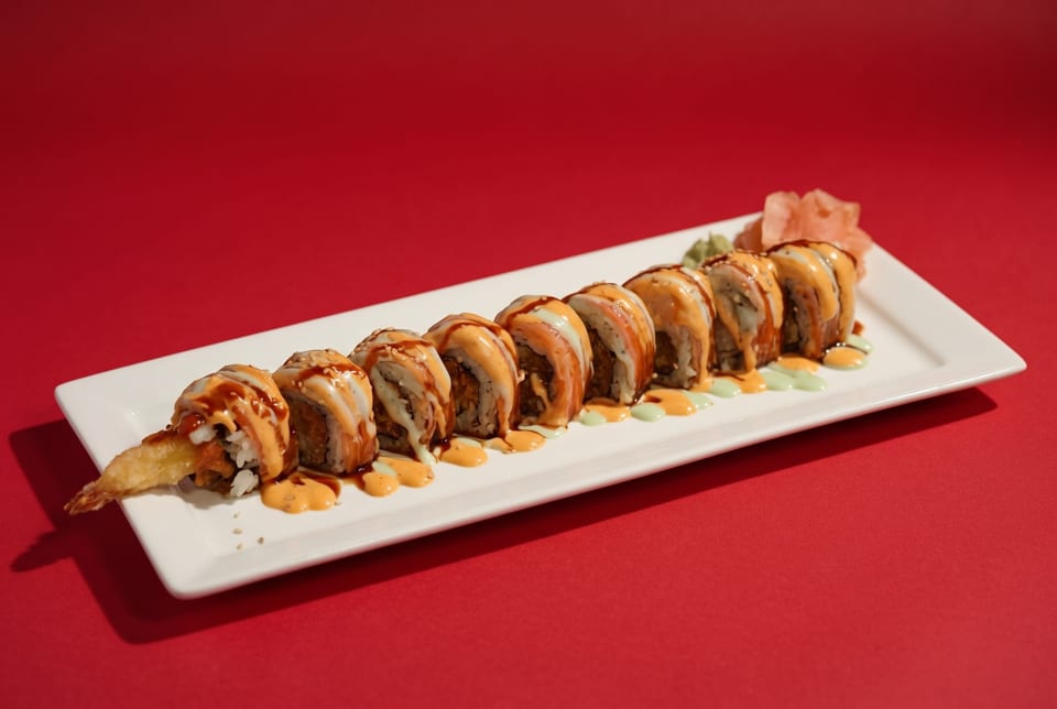 A white rectangular plate holds a row of sushi rolls with sauces and pickled ginger on a red background.